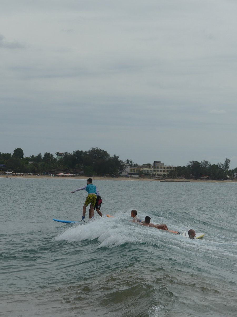 Vitus riding a wave with other surfers paddling behind