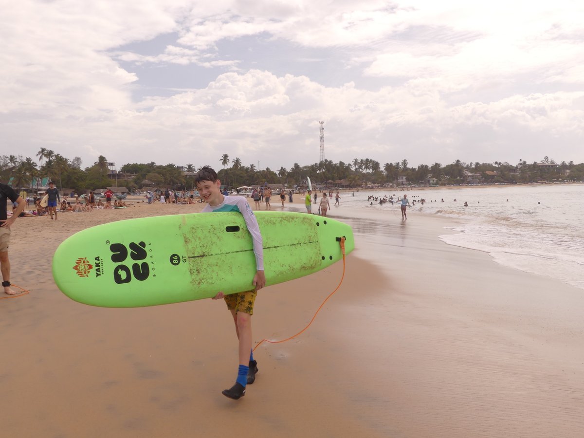 Vitus carrying his green surfboard up the beach, a bootie over his bandaged toe