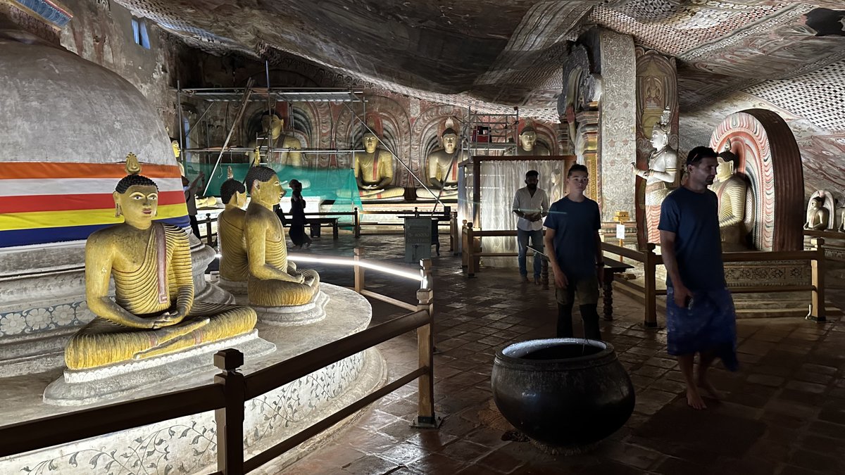 Rows of gilded seated Buddhas along a painted cave hall at Dambulla, Noah and Jesper walking through
