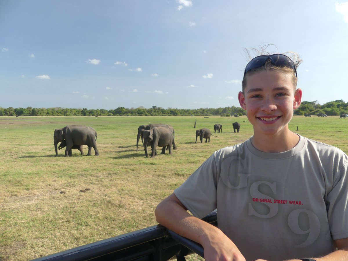 Noah smiling in the jeep with an elephant herd grazing behind him